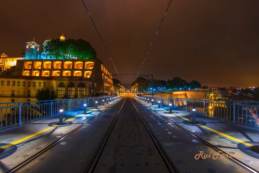 Nighttime view of a railway bridge with illuminated tracks leading towards a historic building and a tower.