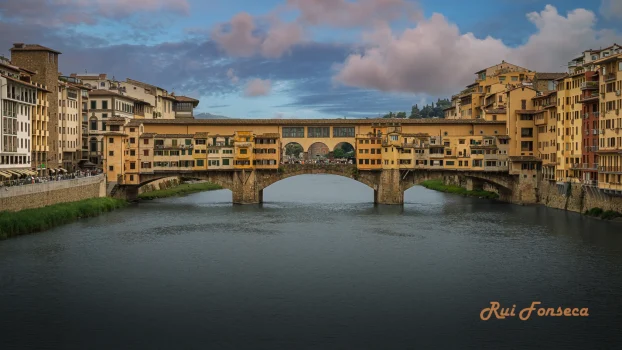 A picturesque view of the Ponte Vecchio bridge in Florence, Italy, with buildings lining the riverbank and a cloudy sky above.