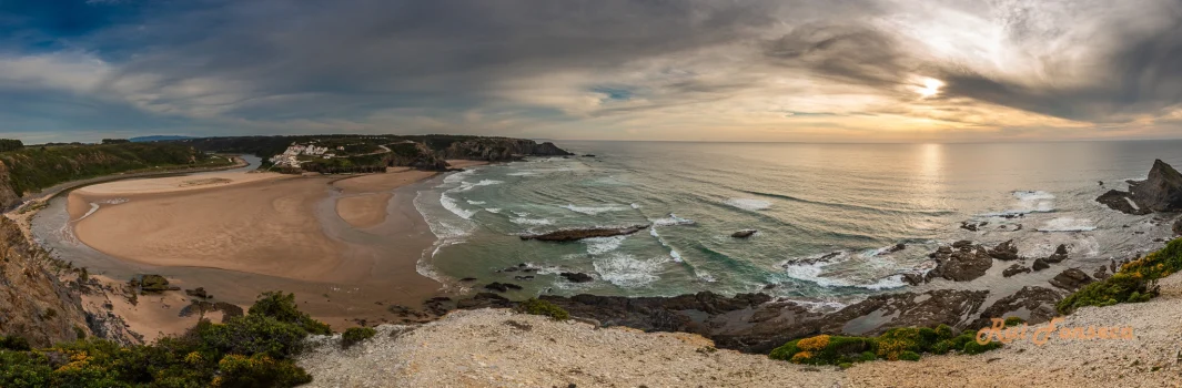 Panoramic view of a coastal landscape with a sandy beach, rocky cliffs, and a cloudy sky with a break revealing sunlight over the ocean.