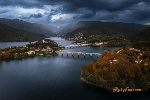 Aerial view of a lake surrounded by mountains, with a stone bridge spanning the water. The sky is overcast with dramatic clouds.