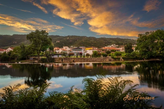 Panoramic view of a village reflected in a calm lake at sunset, with lush vegetation in the foreground.
