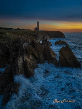 Aerial view of a rugged coastline with crashing waves, a lighthouse in the distance, and a clear sky.