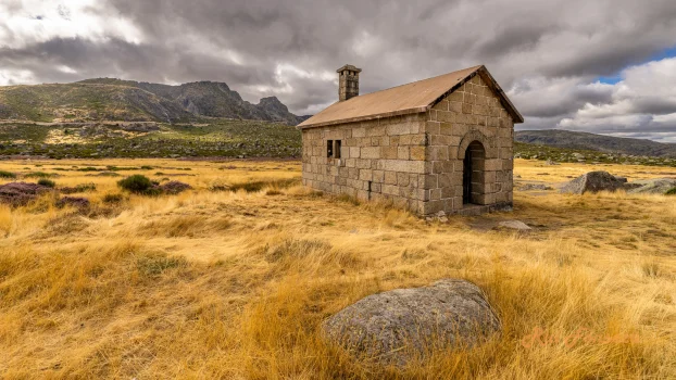 A solitary stone building stands in a vast, golden field under a dramatic, cloudy sky, with mountains visible in the distance.