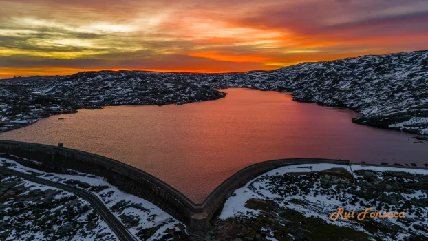 Aerial view of a dam and reservoir surrounded by snow-covered hills under a vibrant sunset.