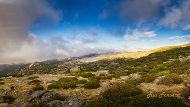 Panoramic view of a mountainous landscape with low-lying vegetation, scattered rocks, and a dramatic sky with clouds and sunlight.