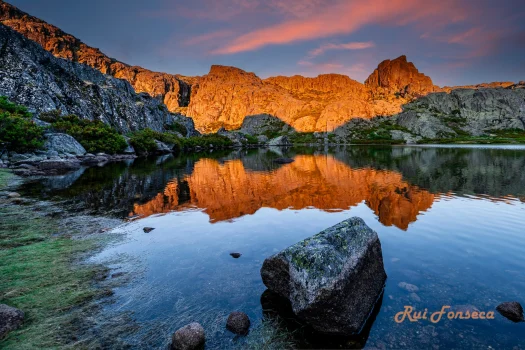 A stunning landscape photograph captures a mountain range illuminated by the golden light of sunrise, reflected in a calm lake. A large rock sits prominently in the foreground.