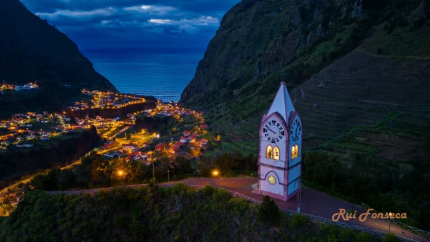 Aerial view of a clock tower in a town nestled between mountains and the ocean at dusk.