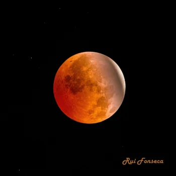 A photograph of a total lunar eclipse, with the moon appearing a deep reddish-orange against a black night sky filled with stars.
