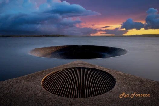 A long exposure photograph of a concrete structure with two circular openings, one above the other, overlooking a calm body of water at twilight. The sky is filled with dramatic clouds in shades of purple, pink, and orange. The foreground is dark, emphasizing the structure and the water's reflection.