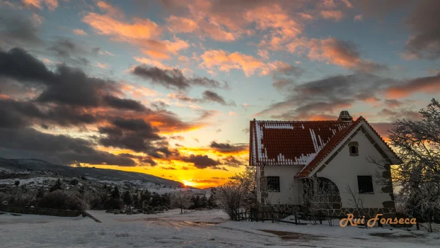 A white building with a red tile roof sits in a snowy landscape under a dramatic sunset sky. The foreground is dominated by snow-covered ground and sparse, snow-laden trees. In the background, rolling hills are visible.