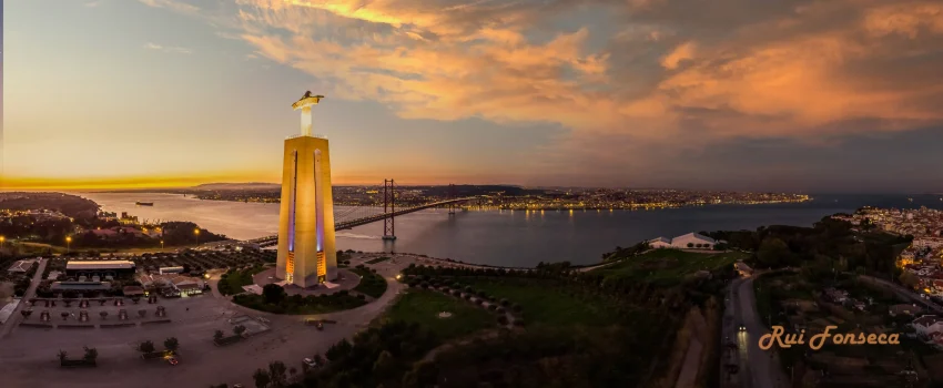 Aerial view of the Monument to the Discoveries in Lisbon, Portugal, at dusk with a bridge and cityscape in the background.