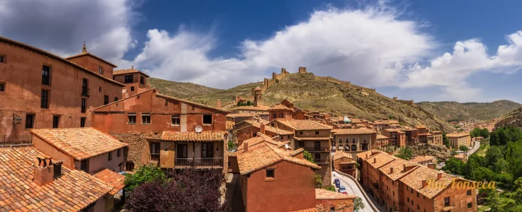 Panoramic view of a historic town with red-tiled roofs, a hilltop castle, and a bright blue sky with scattered clouds.