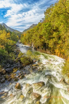 A scenic view of a fast-flowing river surrounded by lush autumn foliage and mountains under a partly cloudy sky.